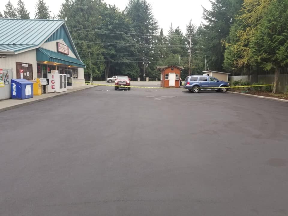 A freshly sealed black parking lot beside a corner grocery store, cars parked at the far edge under grey Pacific Northwest sky.