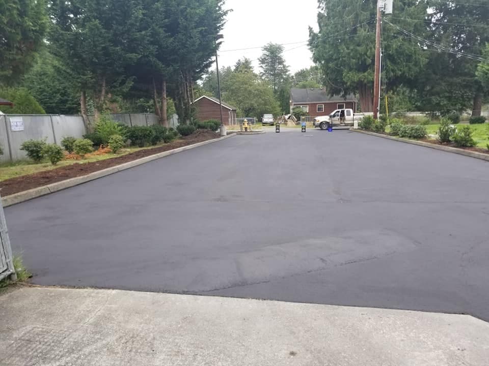 A freshly sealed black asphalt driveway beside a house, fresh landscaping along one edge and a work truck parked at the far end.