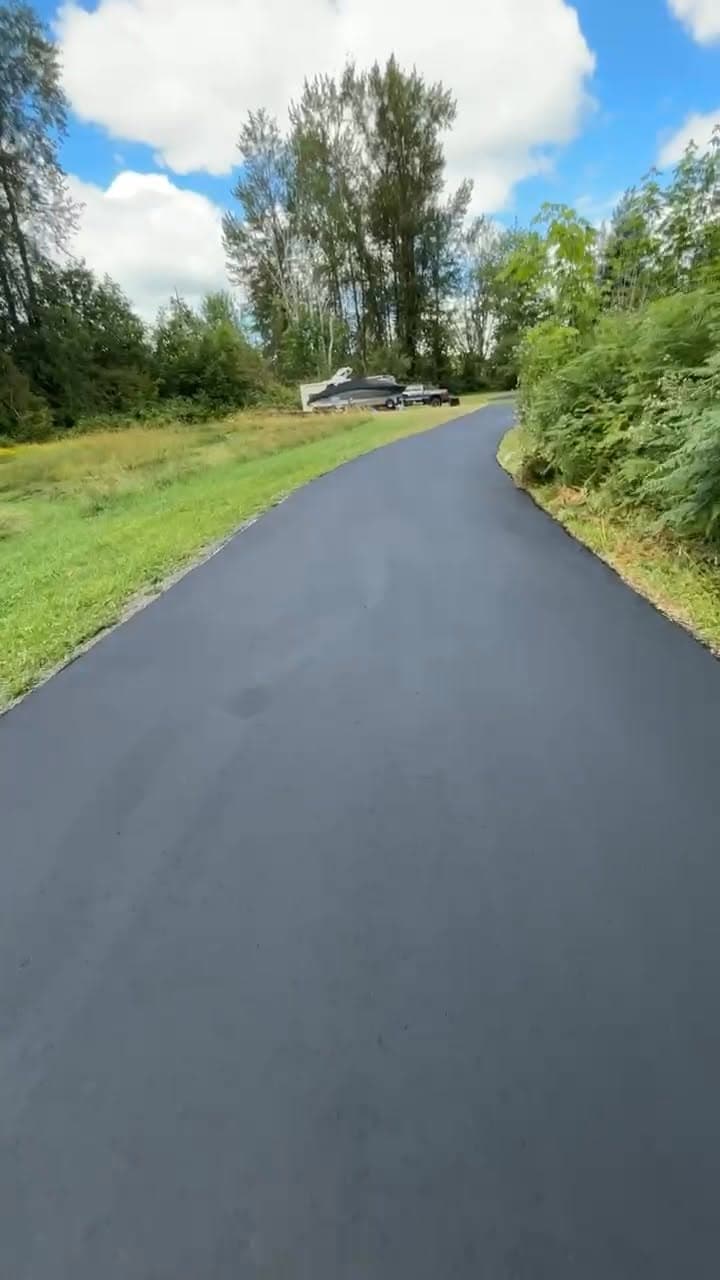 A freshly paved winding asphalt driveway running through a green Marysville property under a blue sky, a boat on a trailer parked off to the side.